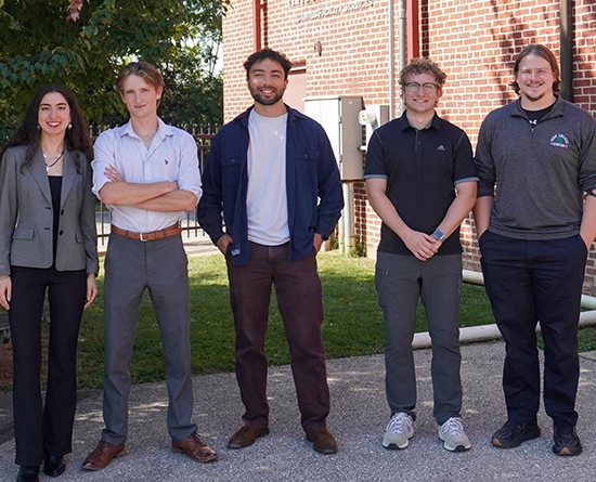 Five students, four men and one woman, standing outside in front of a brick building