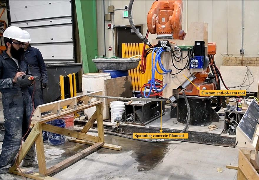 Worker in safety gear operates a robotic arm with a custom end-of-arm tool, extruding a spanning filament of concrete in an industrial workshop.